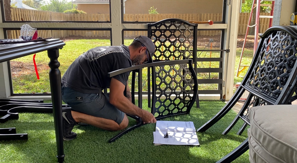 A man holding a patio chair part while kneeling. A man holding a patio chair part while kneeling.