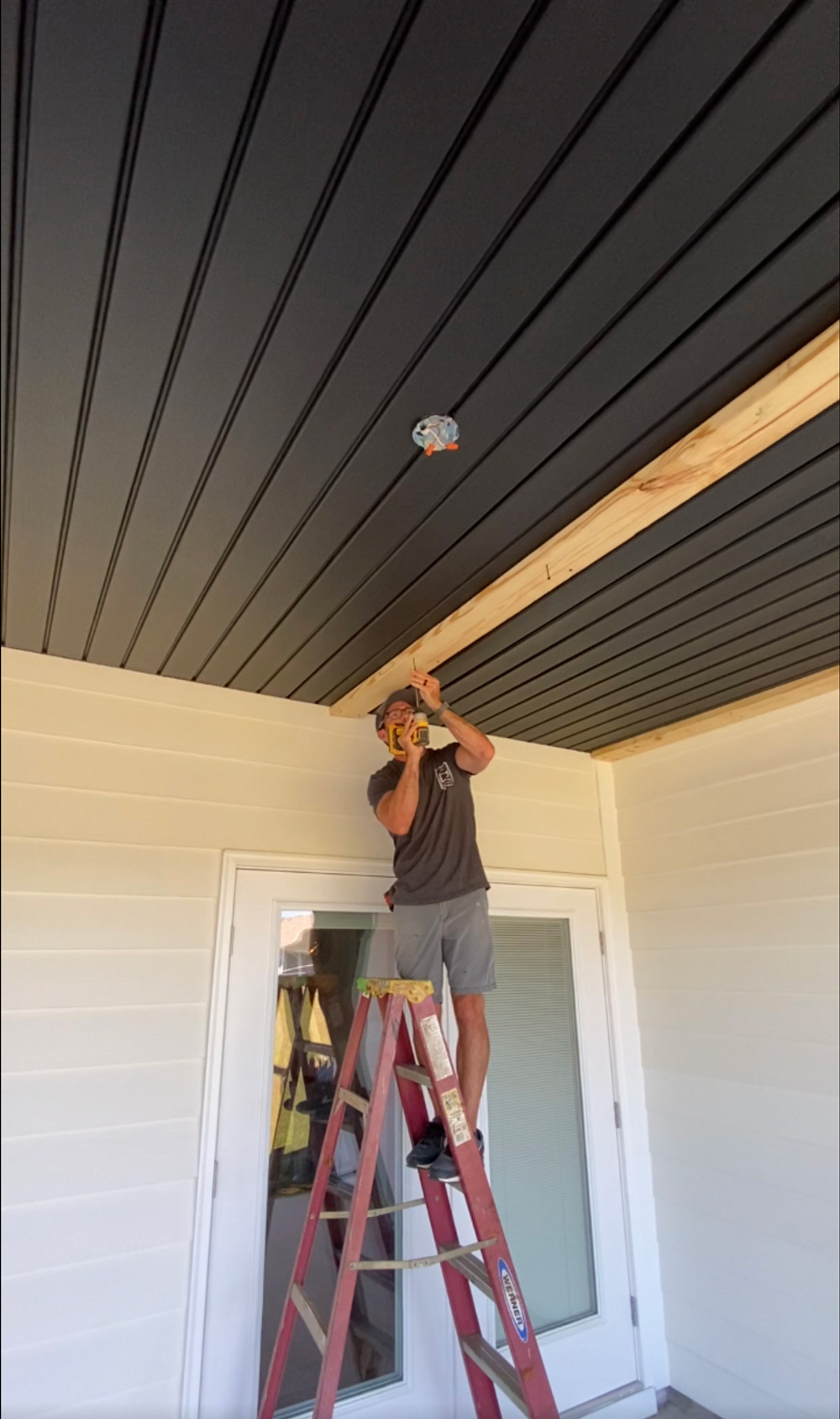 A man standing on a ladder close to the patio door, installing wooden ceiling beams. A man standing on a ladder close to the patio door, installing wooden ceiling beams.