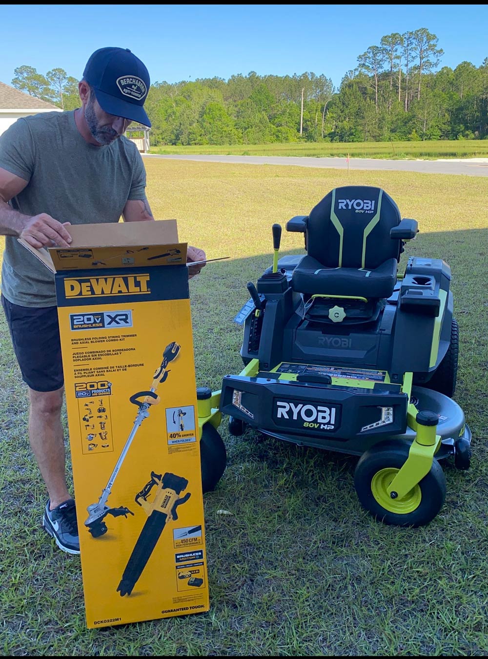 A man holding a DeWalt trimmer box, while standing next to a Ryobi lawnmower. A man holding a DeWalt trimmer box, while standing next to a Ryobi lawnmower.