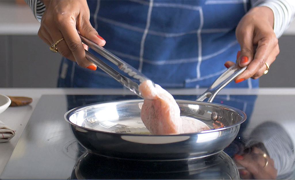 A woman cooks chicken in a Stainless steel pan. 