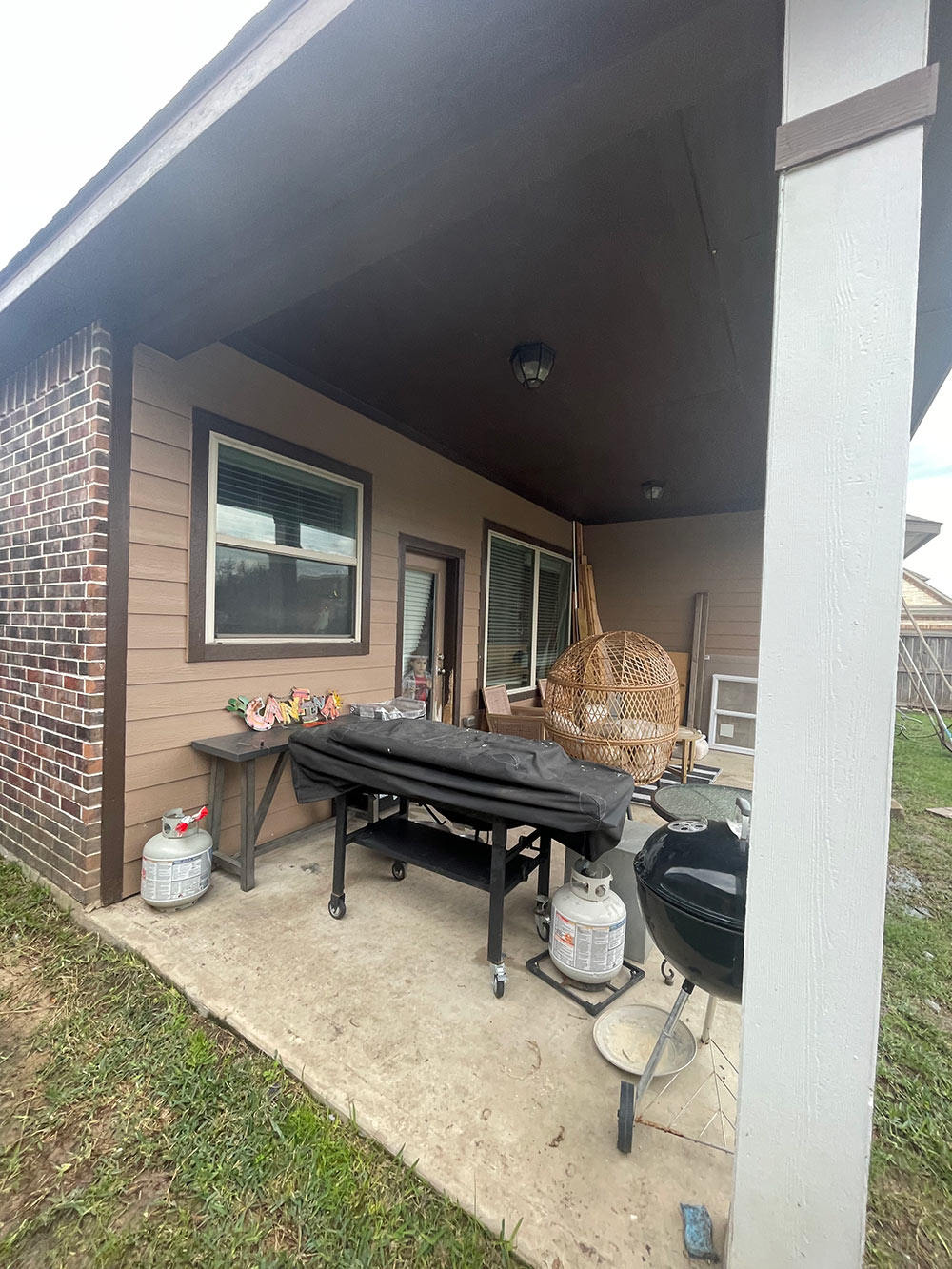 A cluttered back patio with a window, two grills and two propane tanks. A cluttered back patio with a window, two grills and two propane tanks.