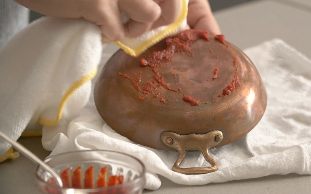 A person rubs tomato paste on the bottom of a copper pan.