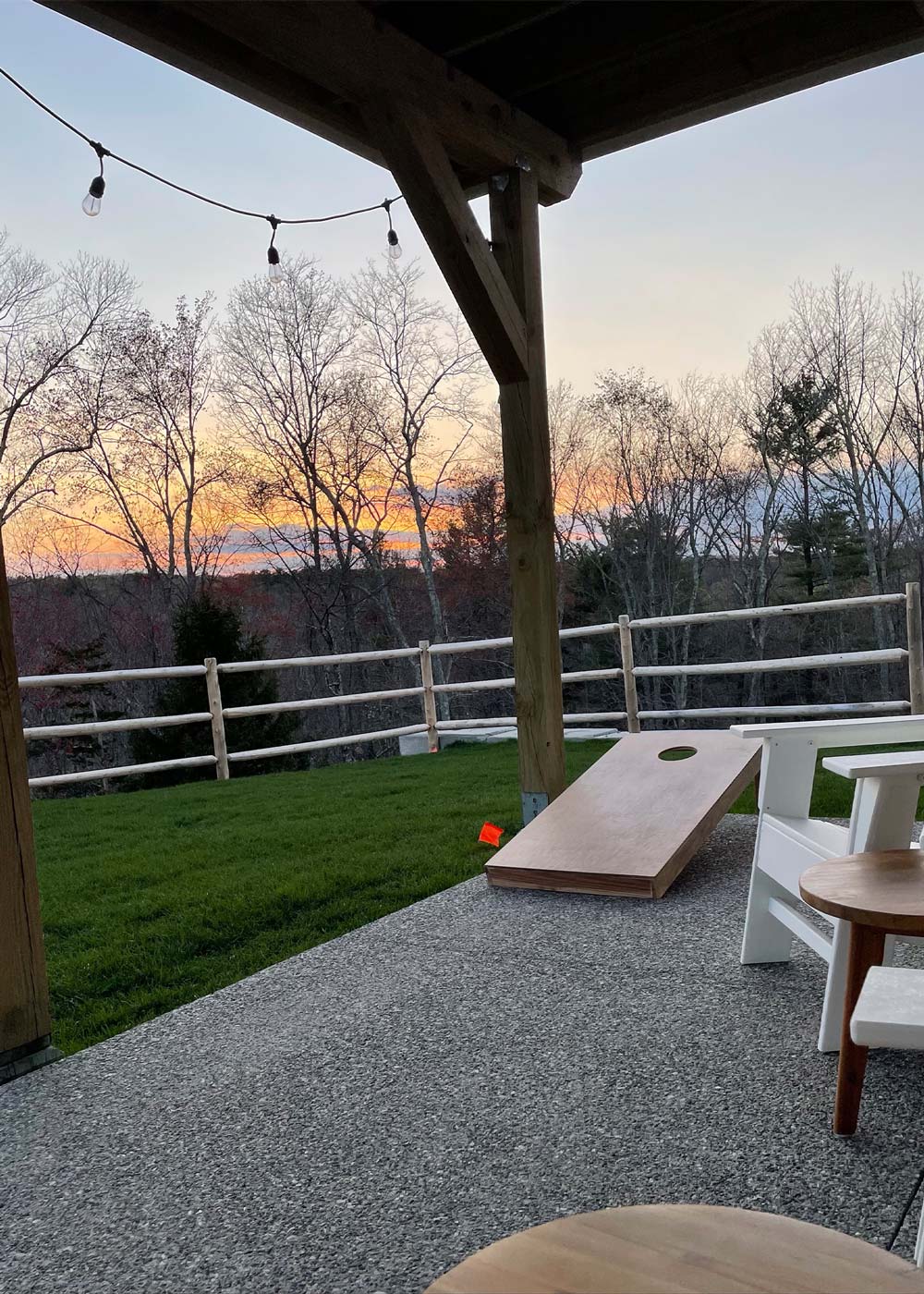 A backyard patio with white lawn chairs and wooden corn hole game, with a sunset in the background