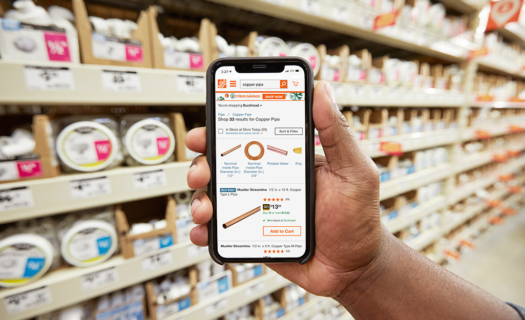 A person holds a smartphone displaying a Home Depot product page while on an aisle in the store.