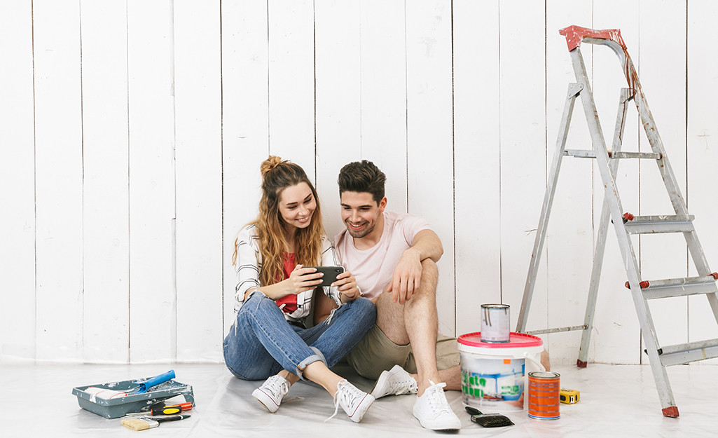 A woman and a man look at a smartphone as they sit on floor next to ladder and paint supplies.