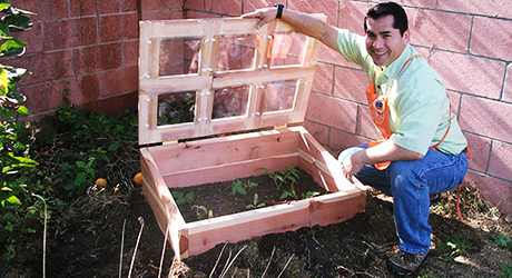 A person kneels next to a cold frame and holds it open.