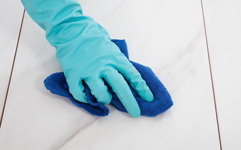 A person's hand wearing gloves and using a cloth to wipe a tile clean.