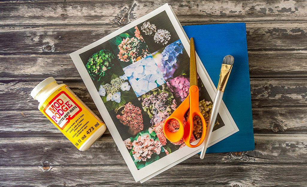 Photos printed on paper, decoupage glue, scissors, paint brush and a piece of wood on a table.