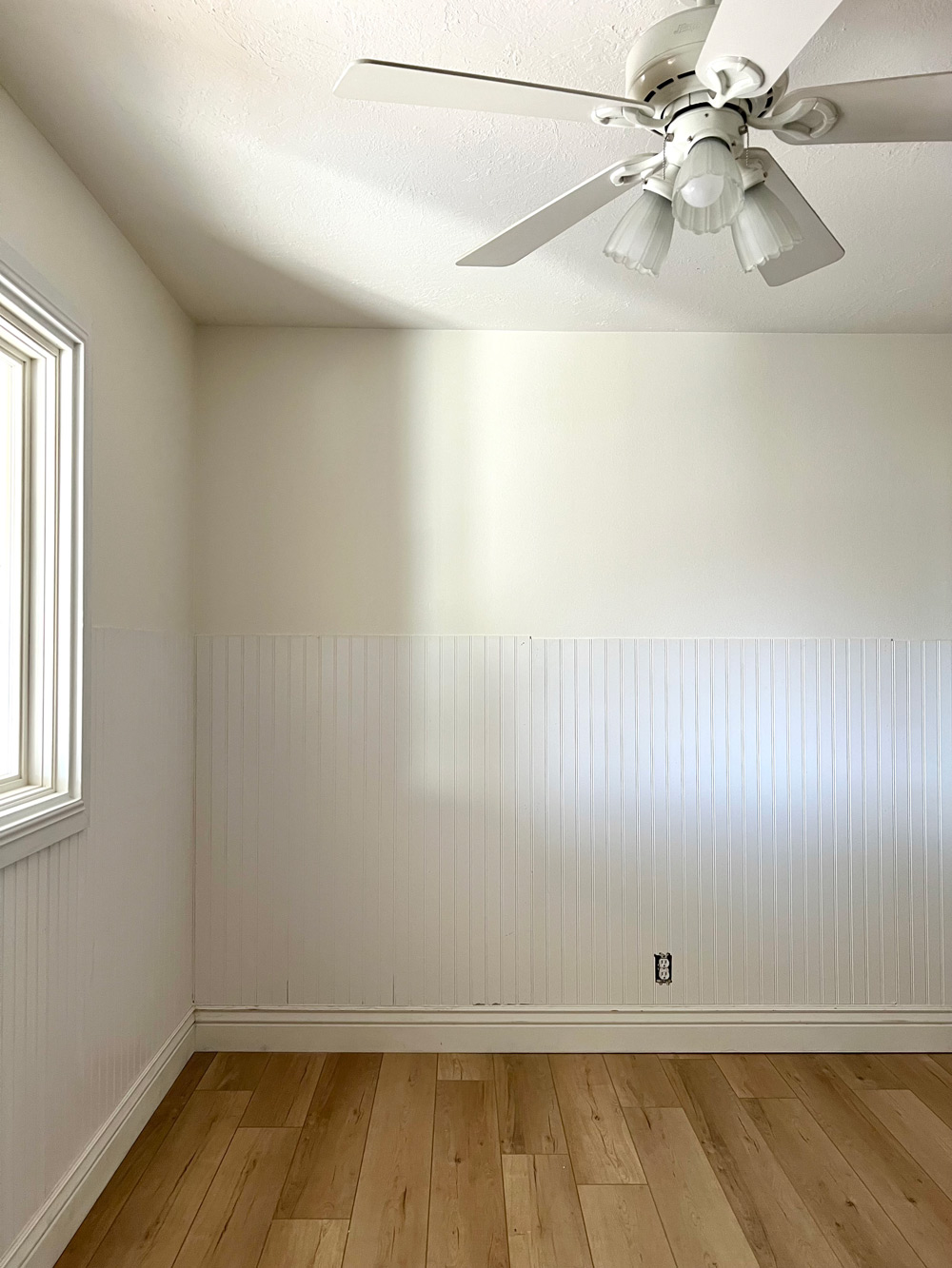 Corner of a beige room with white beadboard and ceiling fan.
