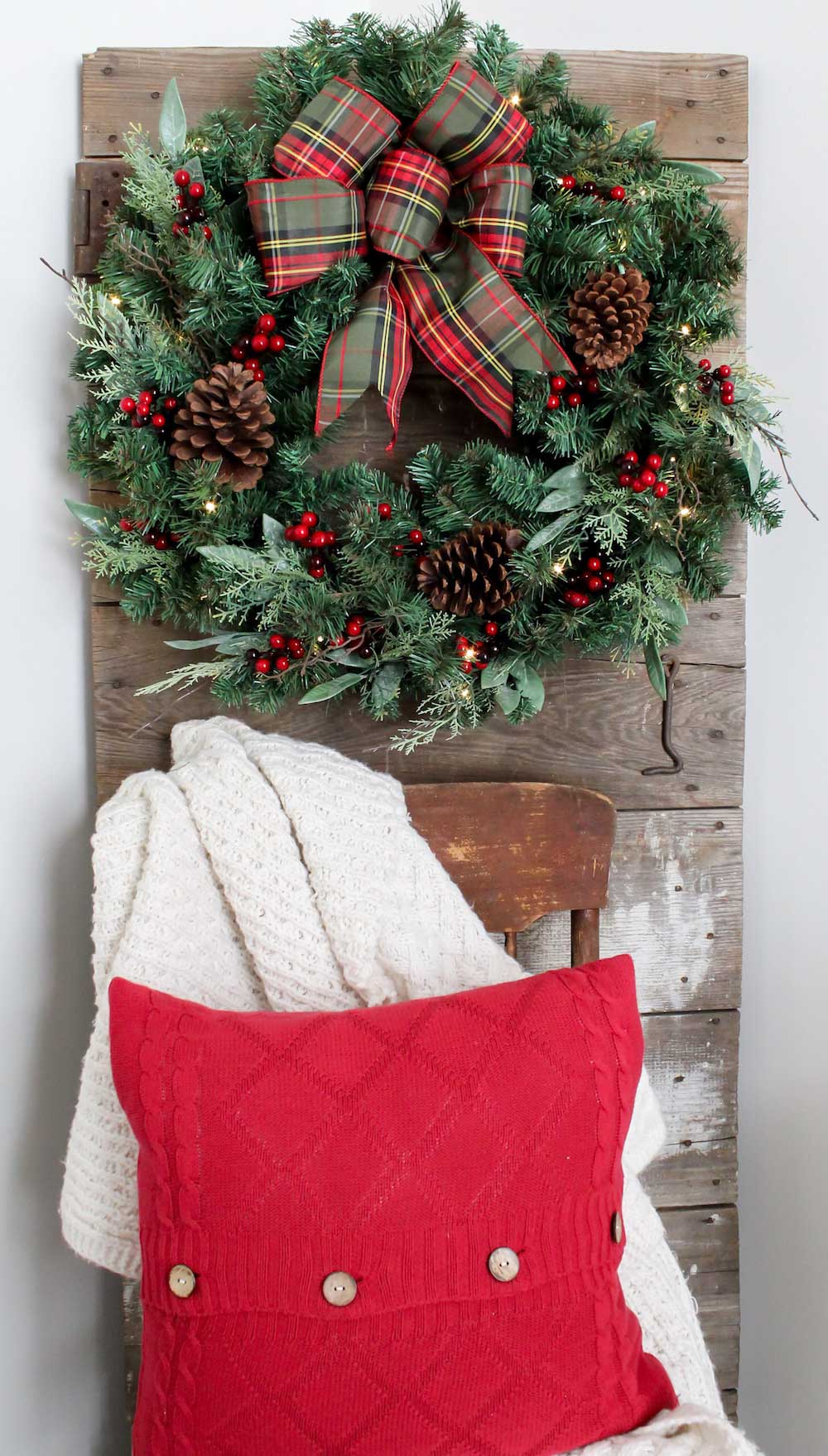Holiday wreath hanging on wooden wall panel with a chair in front covered with a blanket and red pillow
