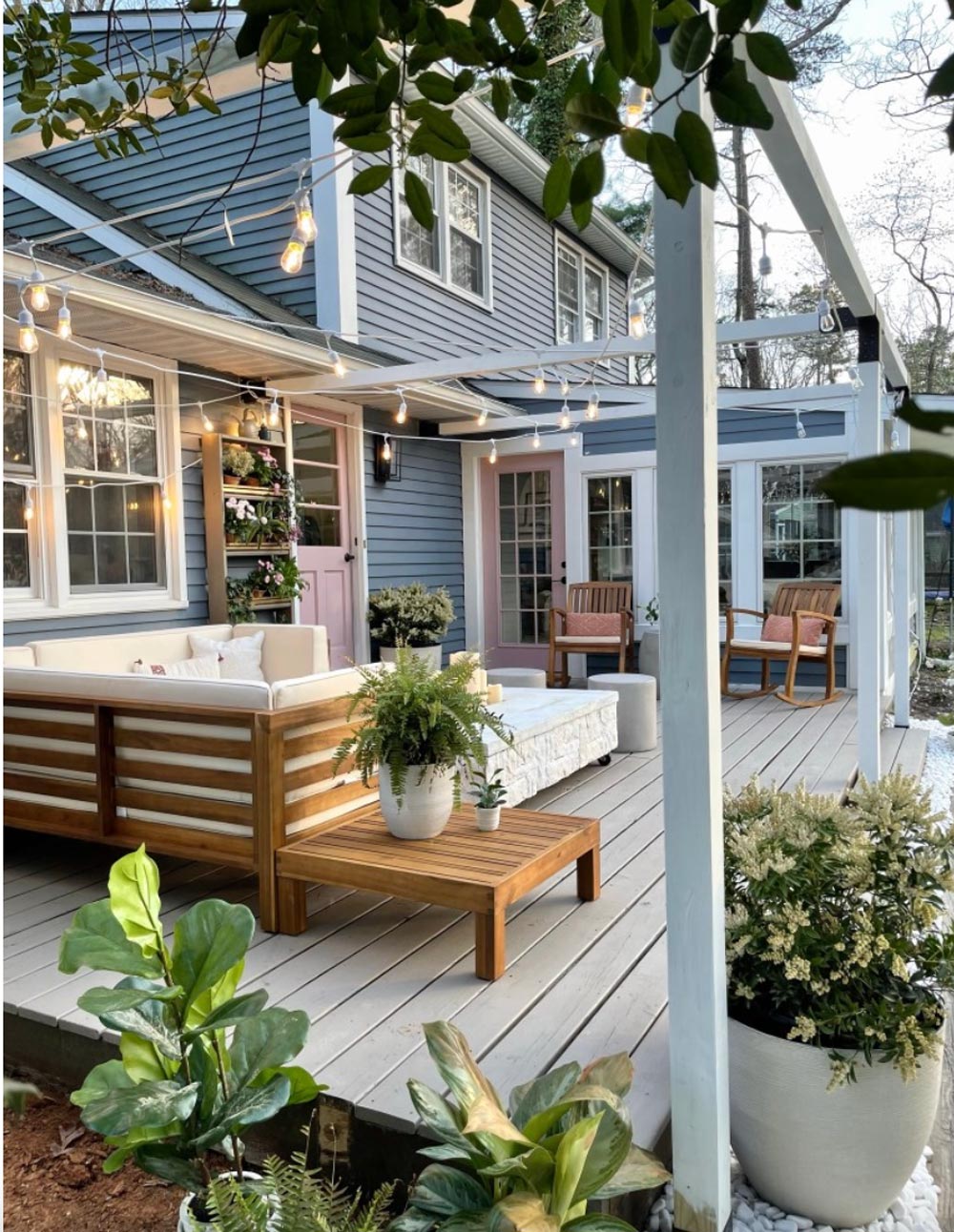 Blue house with updated wood deck with white pergola, pink doors, and white and light wood furniture.