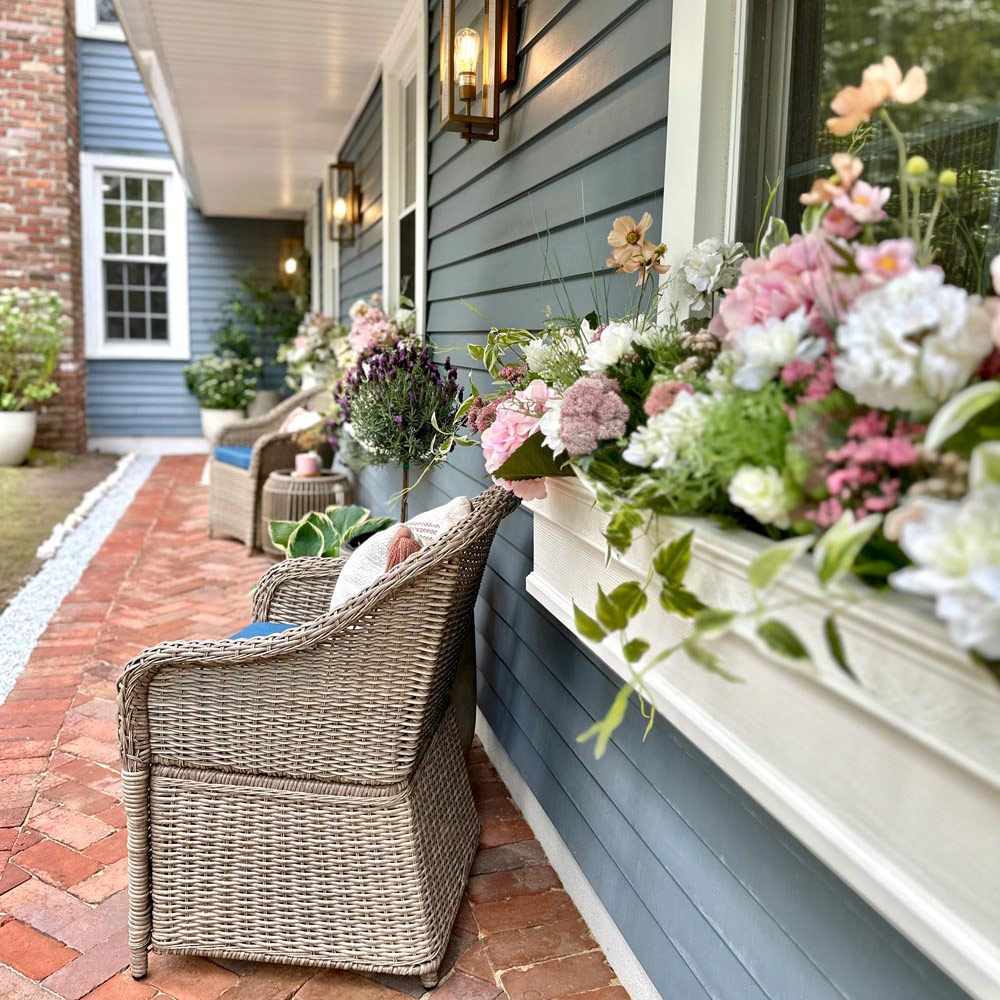 Brick front porch of slate blue house with pink and white flowers in window planters. 