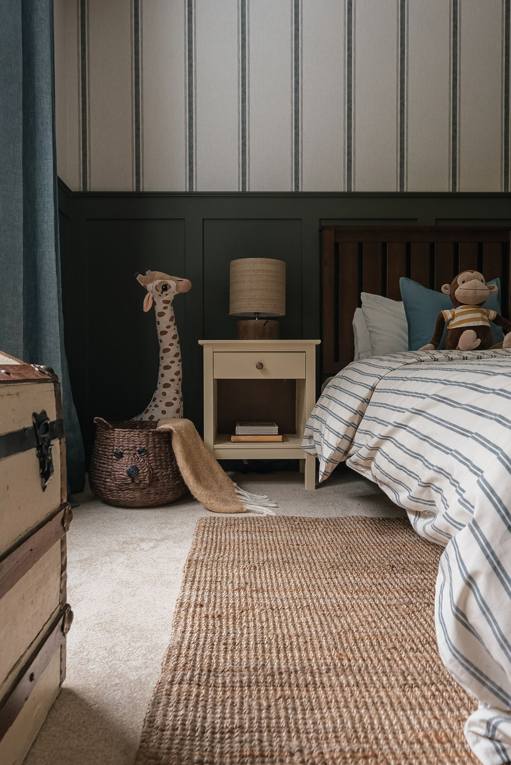 The corner of a child’s bedroom decorated with a teddy bear basket, giraffe toy, nightstand, and the side of a bed.