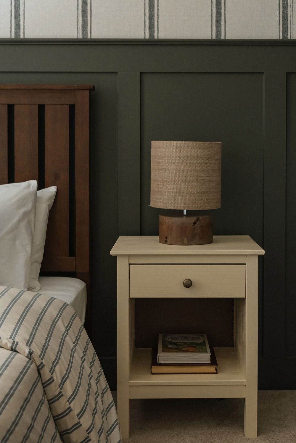 A nightstand with a small lamp, in front of a striped wallpaper, and boarded dark green wall. 
