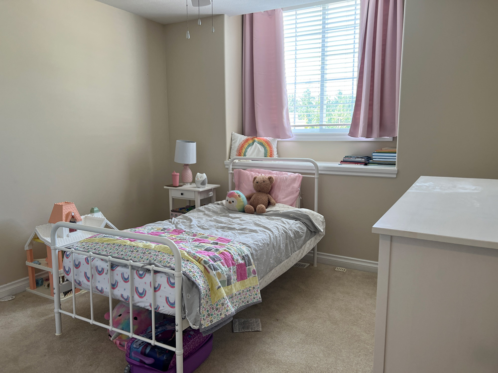 Before photo of a girl’s bedroom with a white frame bed with a teddy bear, and window with pink curtains. Before photo of a girl’s bedroom with a white frame bed with a teddy bear, and window with pink curtains.