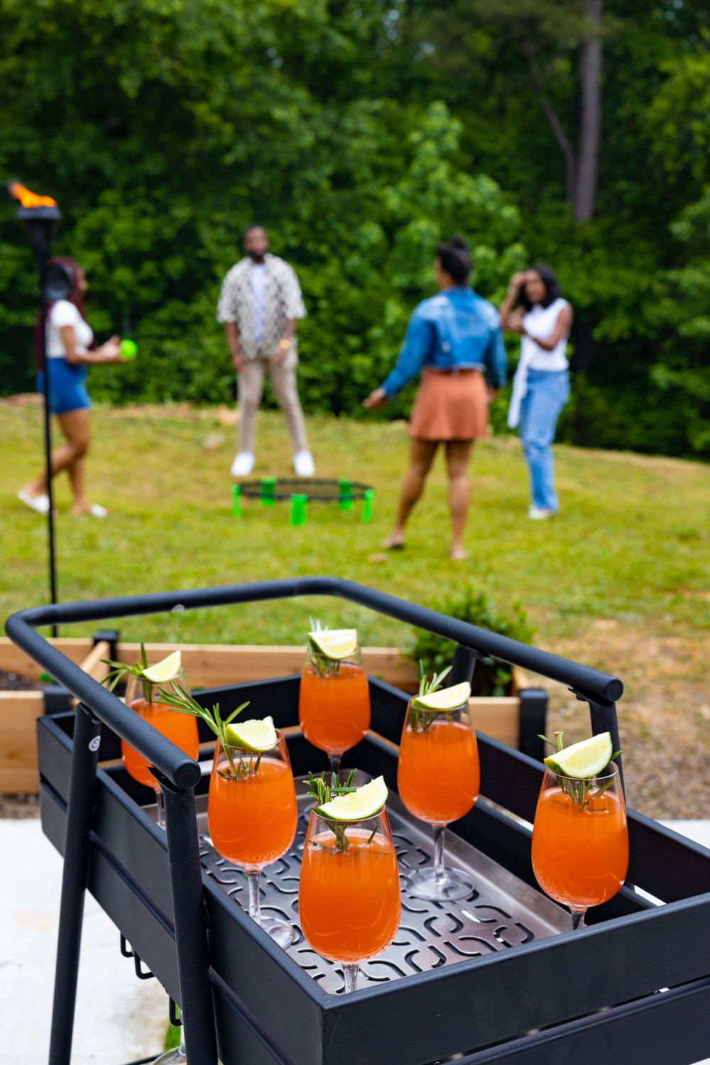 A group of people in the background, and a bar cart with six mocktails in the foreground. A group of people in the background, and a bar cart with six mocktails in the foreground.