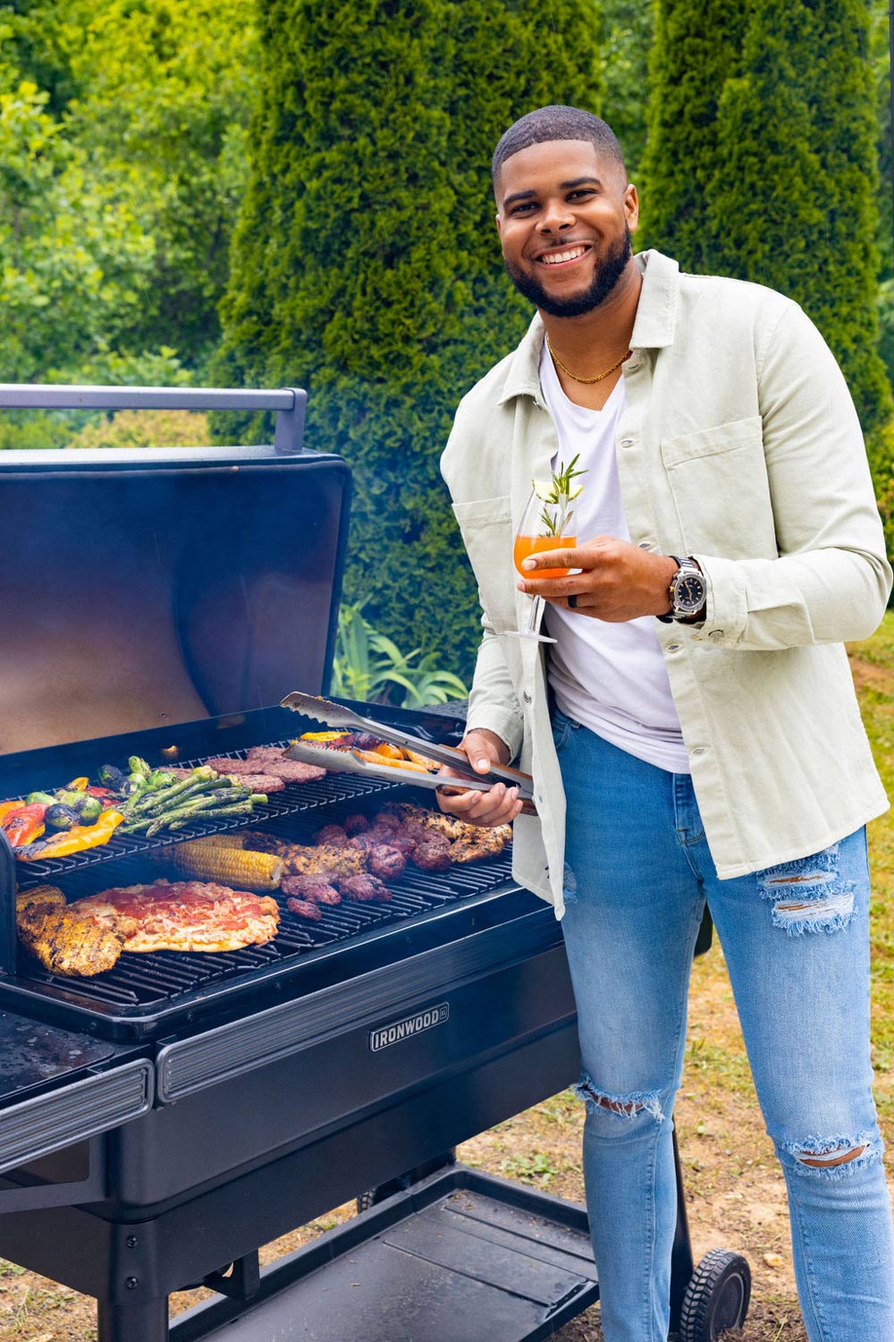 A man smiling and holding a mocktail, next to a grill filled an assortment of foods. A man smiling and holding a mocktail, next to a grill filled an assortment of foods.