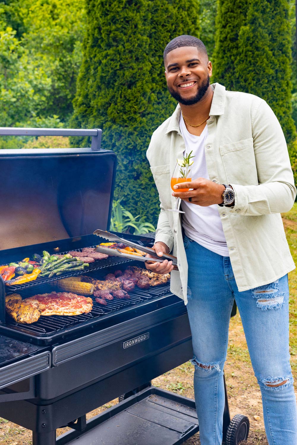 A man smiling and holding a mocktail, next to a grill filled an assortment of foods.