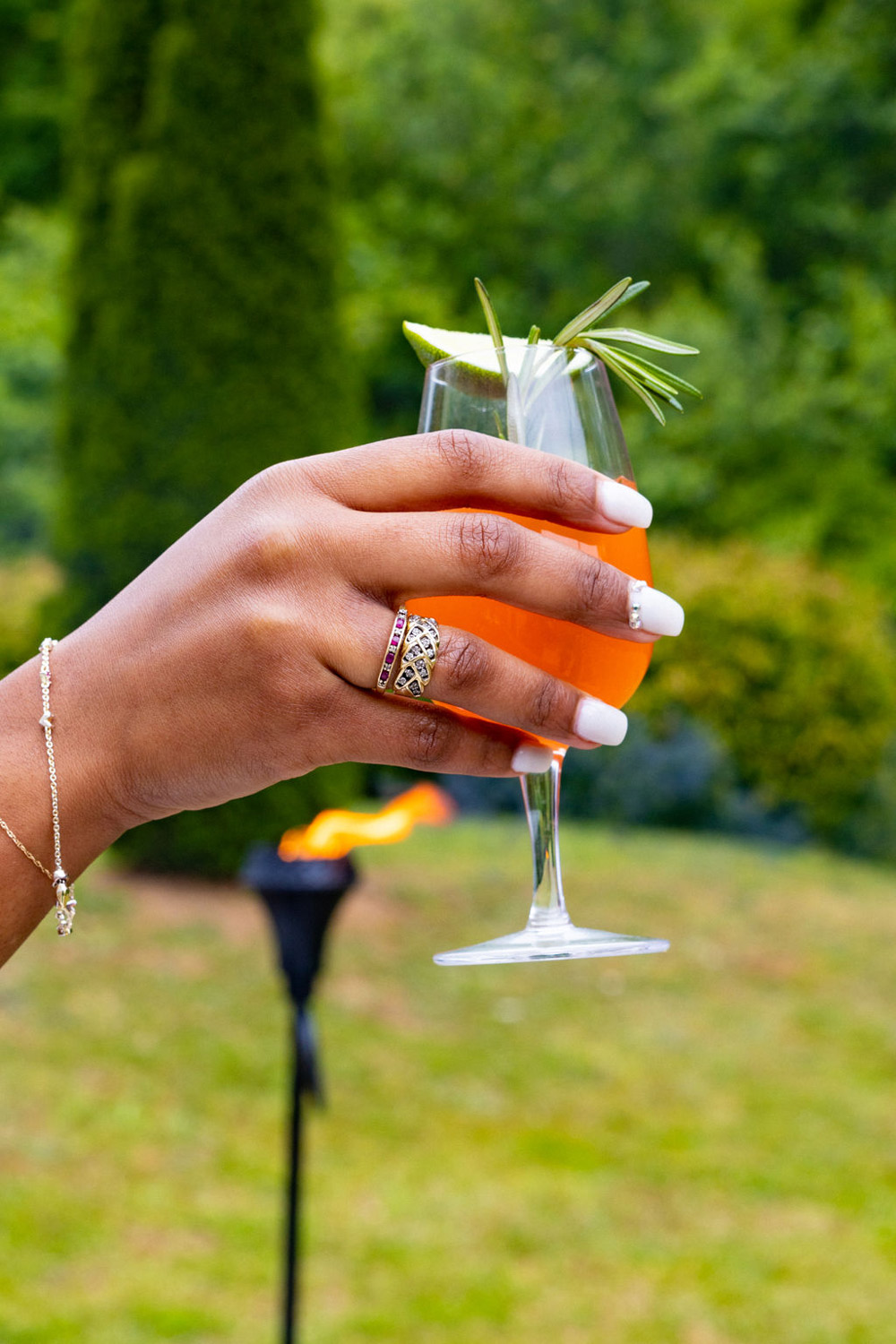 A person wearing a ring and bracelet, holding a garnished mocktail. 