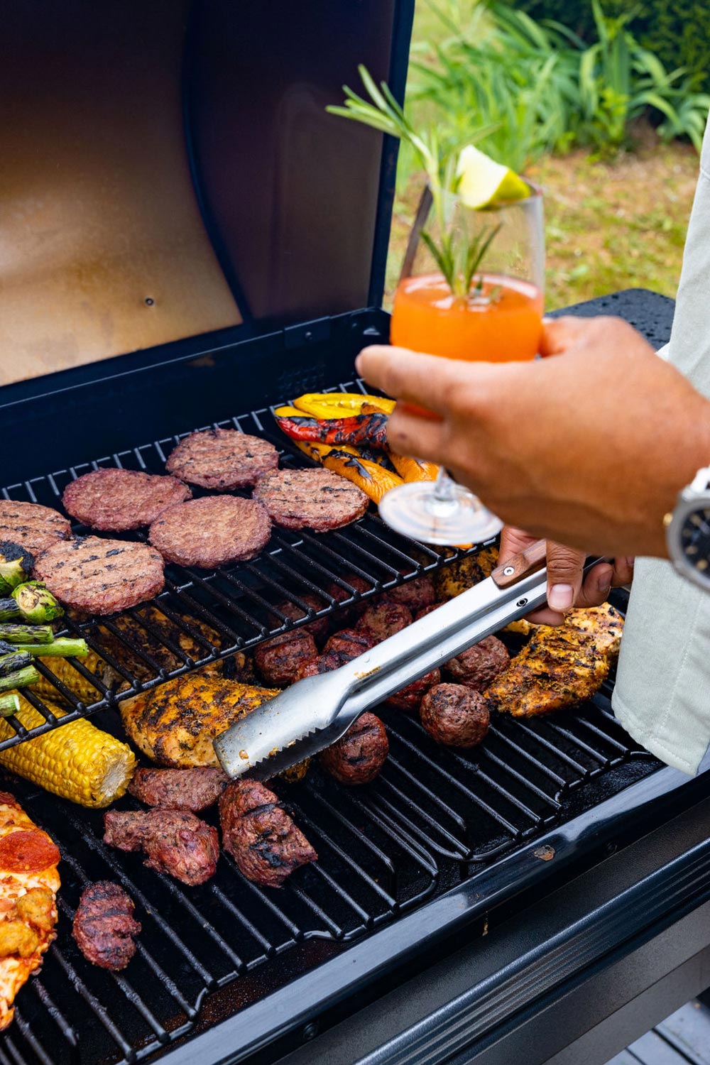A person holding a mocktail while grilling a variety of foods.