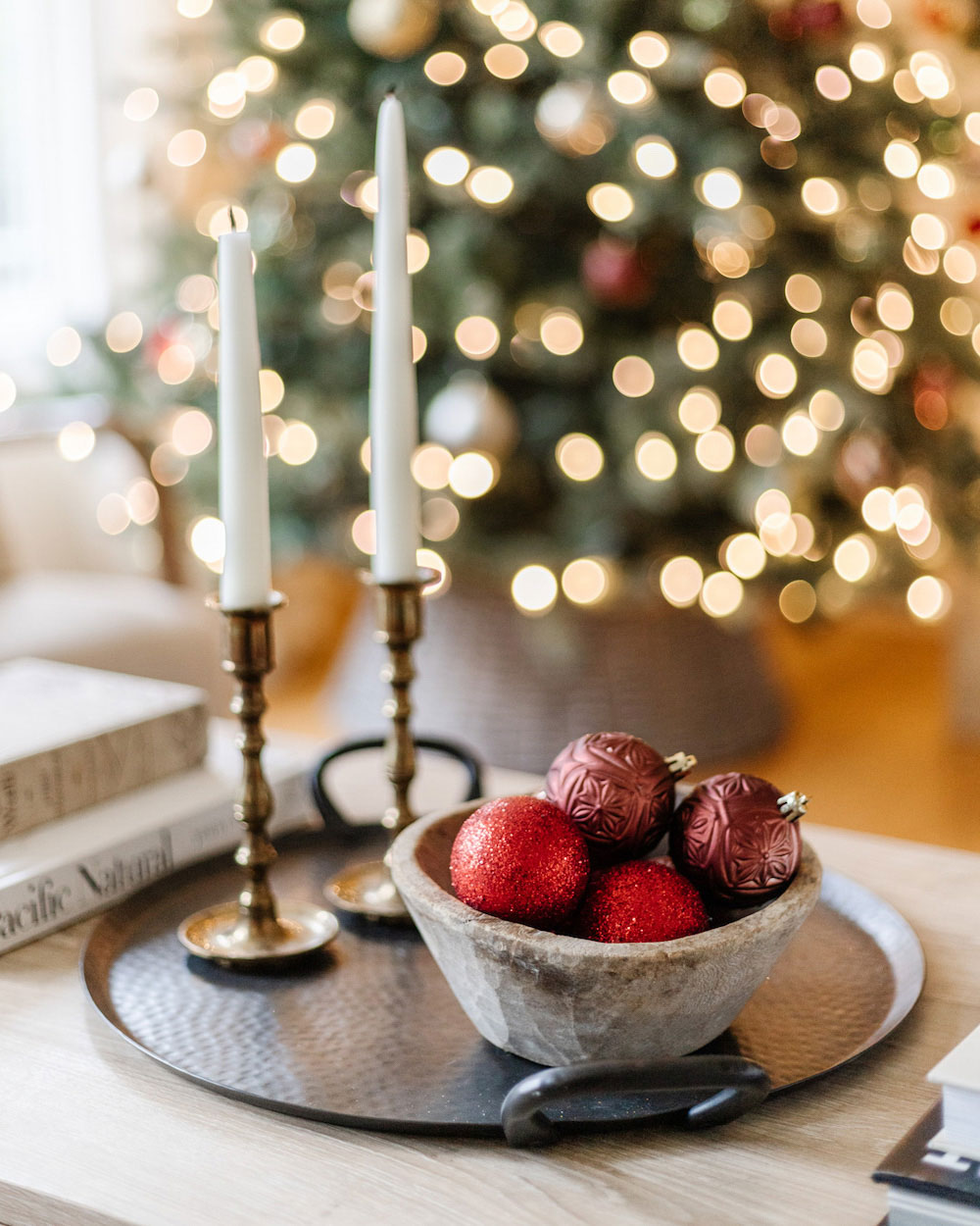 Close up image of a bowl of ornaments and two white candle sticks placed on a coffee table with blurred Christmas lights in the background