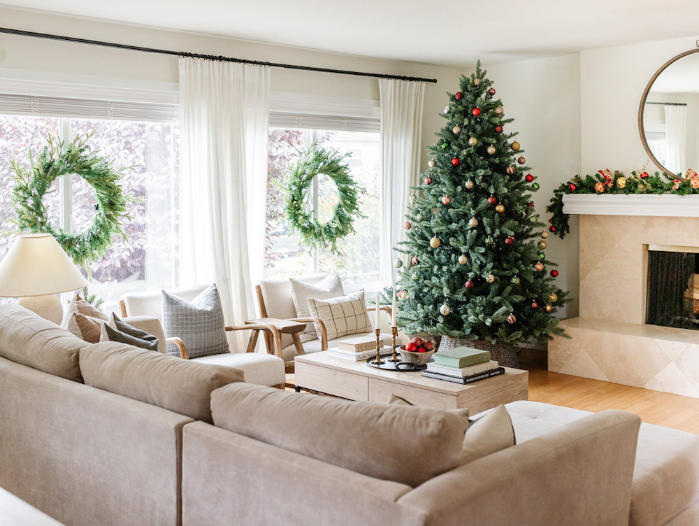 Wide shot of a holiday themed living room that contains beige furniture, a Christmas tree and fireplace in the far back corner and holiday wreaths in the window