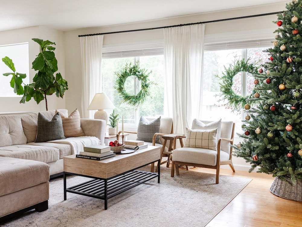 Corner angle of a holiday themed living room with coffee table in the center beige furniture, a Christmas tree in the corner and two wreaths in the back windows