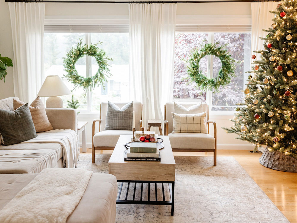 Center shot of a holiday themed living room that contains beige furniture, a coffee table, a Christmas tree in the right corner and wreaths in the back window