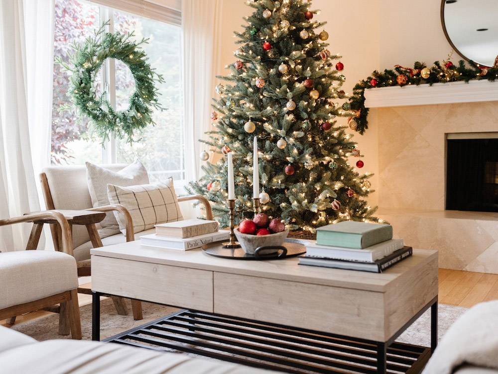 An angled shot of a holiday themed living containing beige furniture, a decorated coffee table, a Christmas tree centered in the background next to a fireplace and wreath
