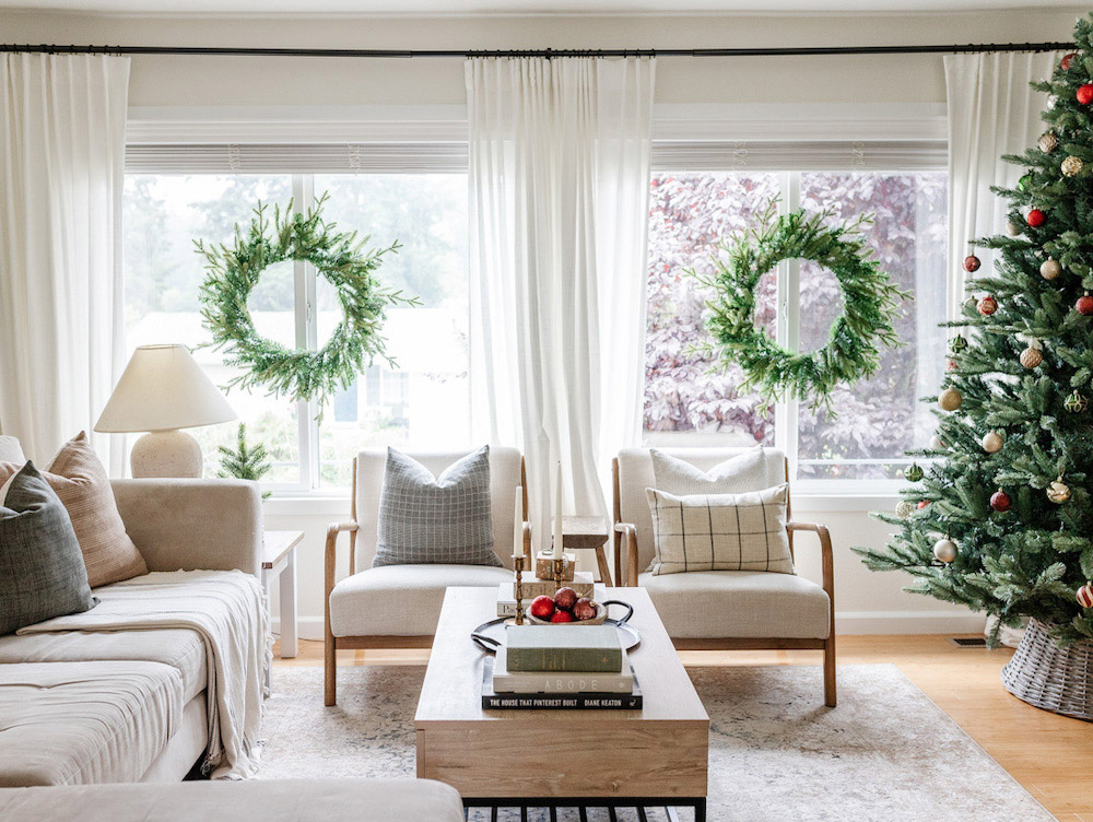 Wide shot of a holiday themed living room that contains a Christmas tree and two wreaths in the back windows