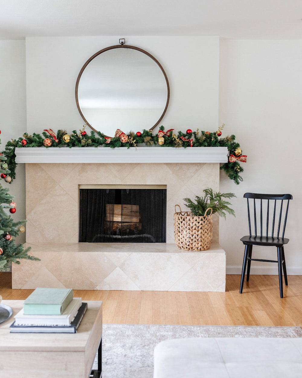 Fireplace topped with holiday garland with a circular mirror on top and a chair beside it