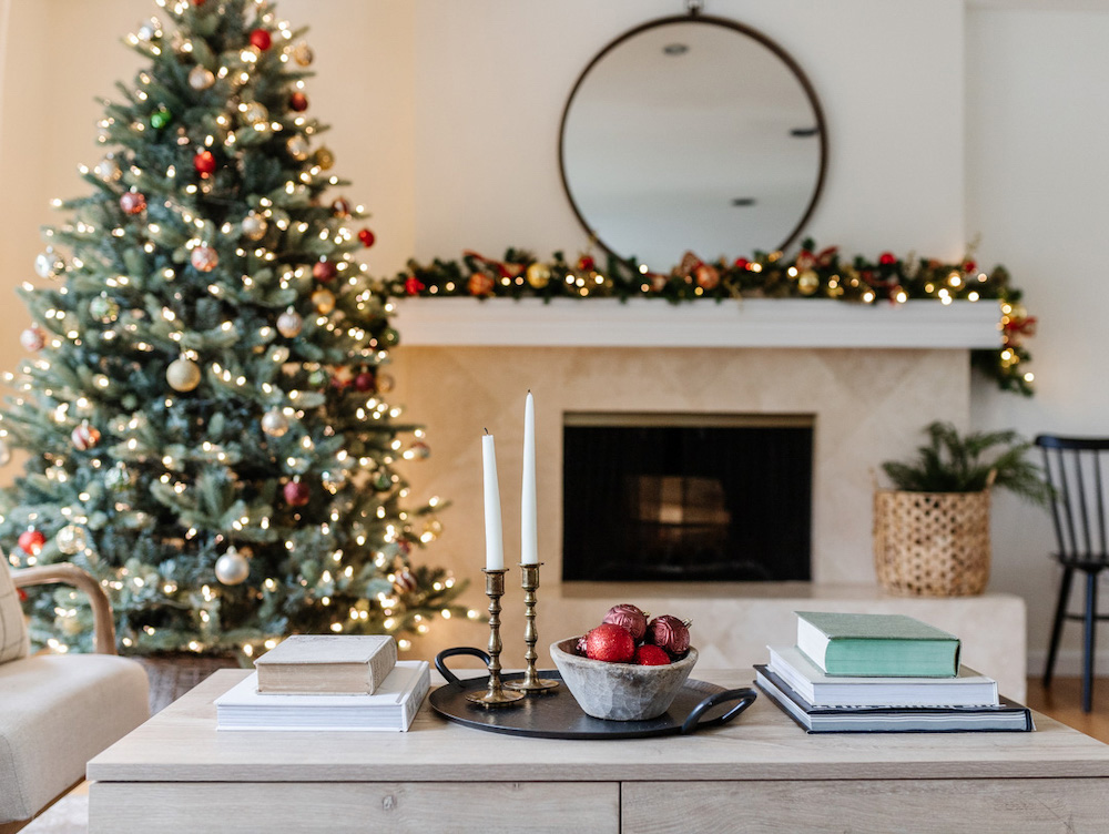 Holiday themed living containing a decorated coffee table, a lit Christmas tree and a fireplace in the background topped with a lit garland and circular mirror 