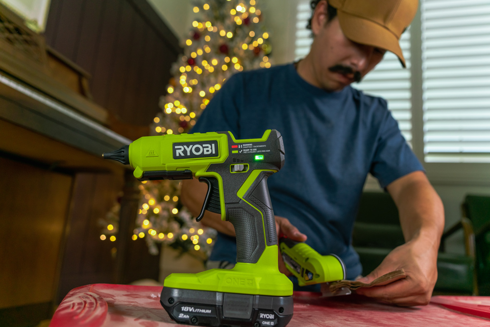 Ryobi 18V hot glue gun focused, with a man using a power cutter to make shapes in a paper bag. Ryobi 18V hot glue gun focused, with a man using a power cutter to make shapes in a paper bag.