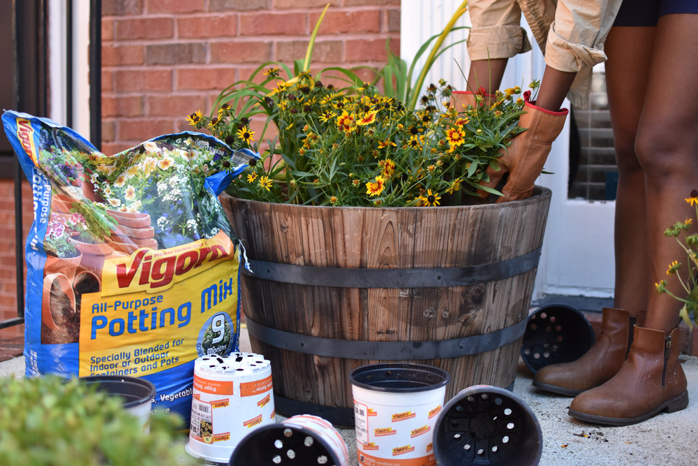 Close up of a woman placing yellow flowers in a decorative wooden flower pot next to a bag of Vigoro potting mix. Close up of a woman placing yellow flowers in a decorative wooden flower pot next to a bag of Vigoro potting mix.