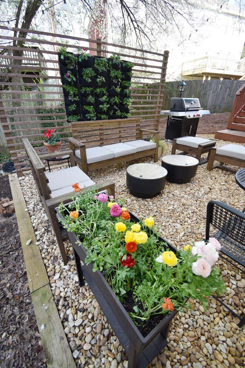 Backyard outdoor patio with wood and canvas outdoor furniture, bed of color flowers, gravel ground, and black grill.