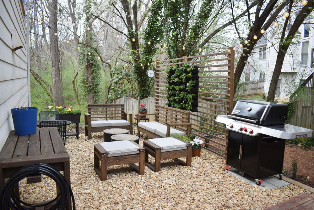 Backyard patio with gravel, string lights, grill, and wooden and canvas patio furniture.
