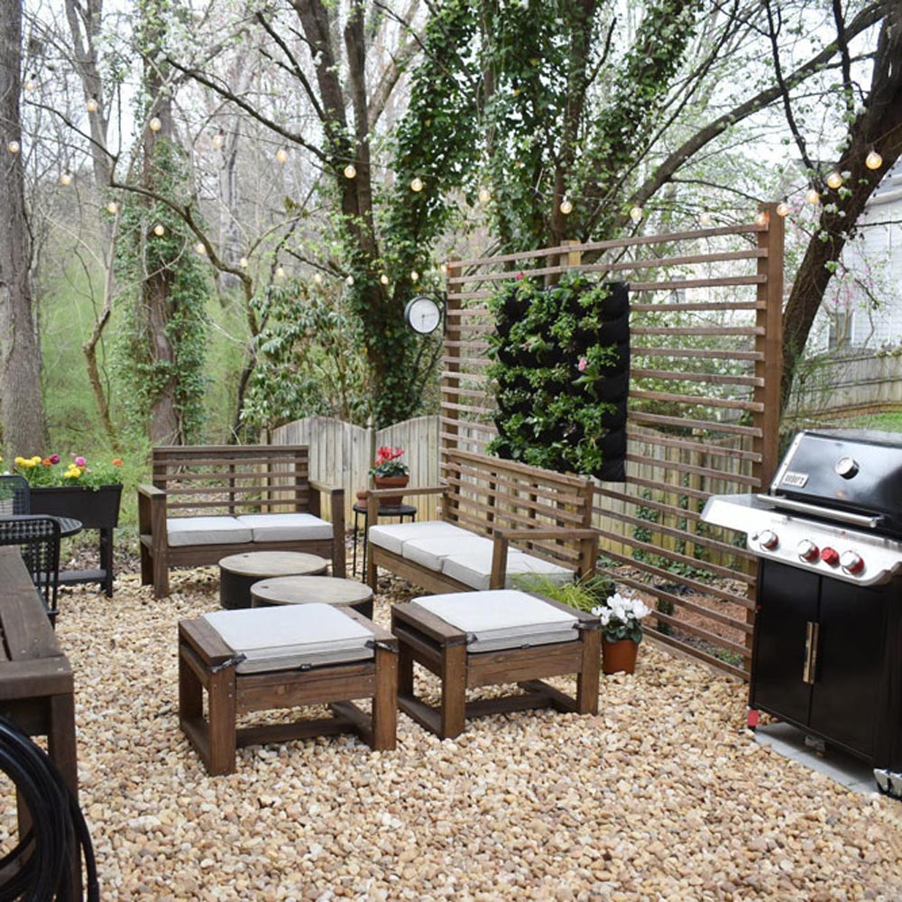 Backyard patio with gravel, string lights, grill, and wooden and canvas patio furniture.