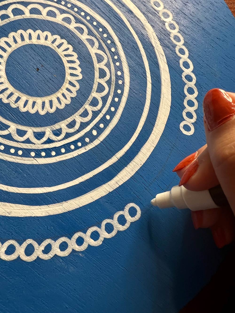 Person making designs on a blue wooden circle with a white paint marker.