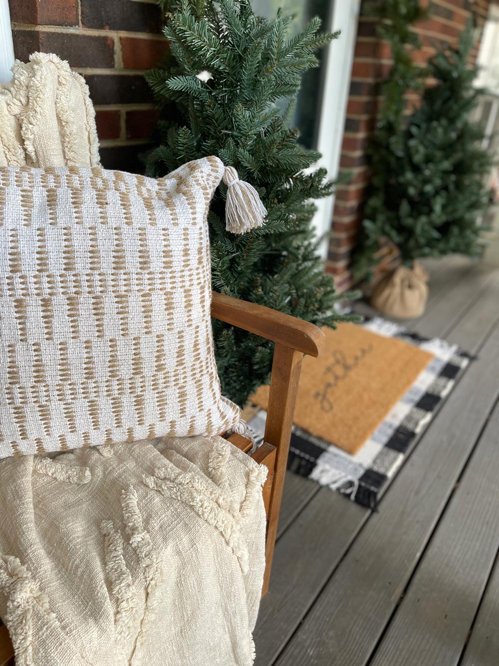 Front porch with mini trees, a “gather” mat, and a bench with a cream-colored throw pillow and blanket.
