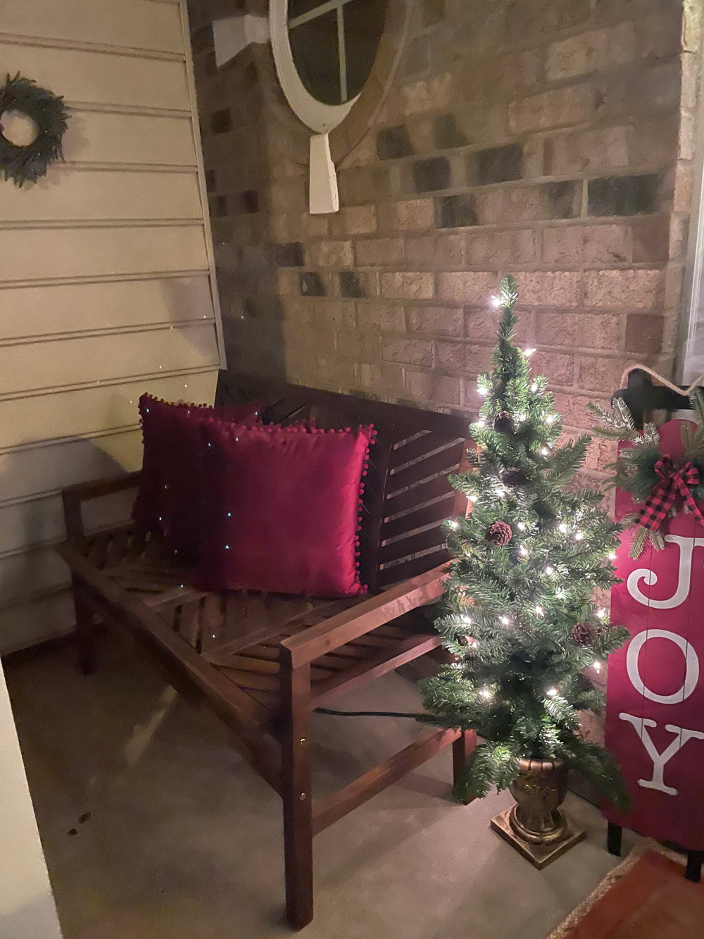 Front porch with a lit mini tree, JOY sign, and burgundy pillows on a bench.