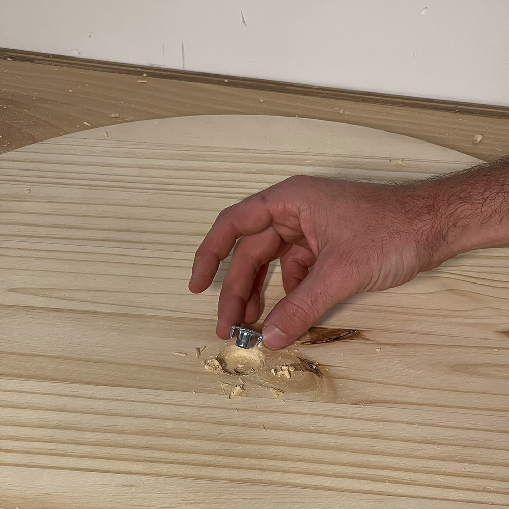 Someone holding a hanger bolt outside of the hole drilled in the wooden circle.