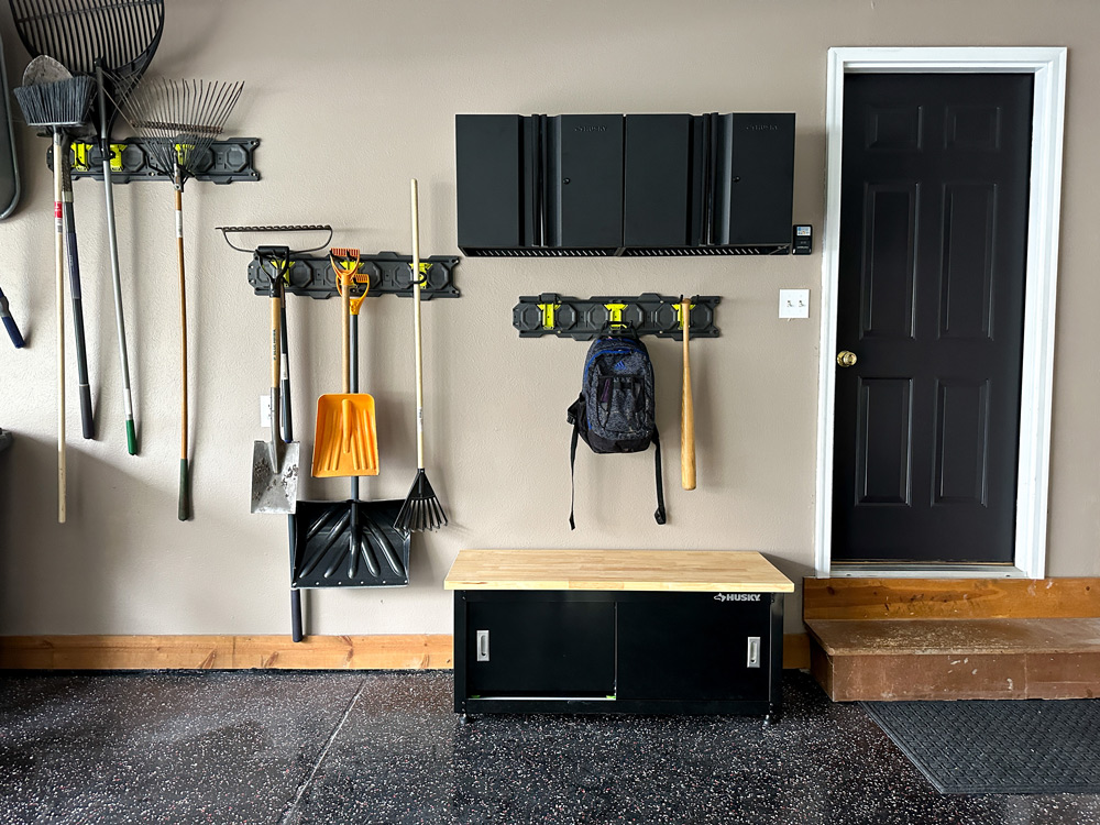 A garage wall with shovels, rakes, a wall storage unit, and a Husky cabinet on the ground.