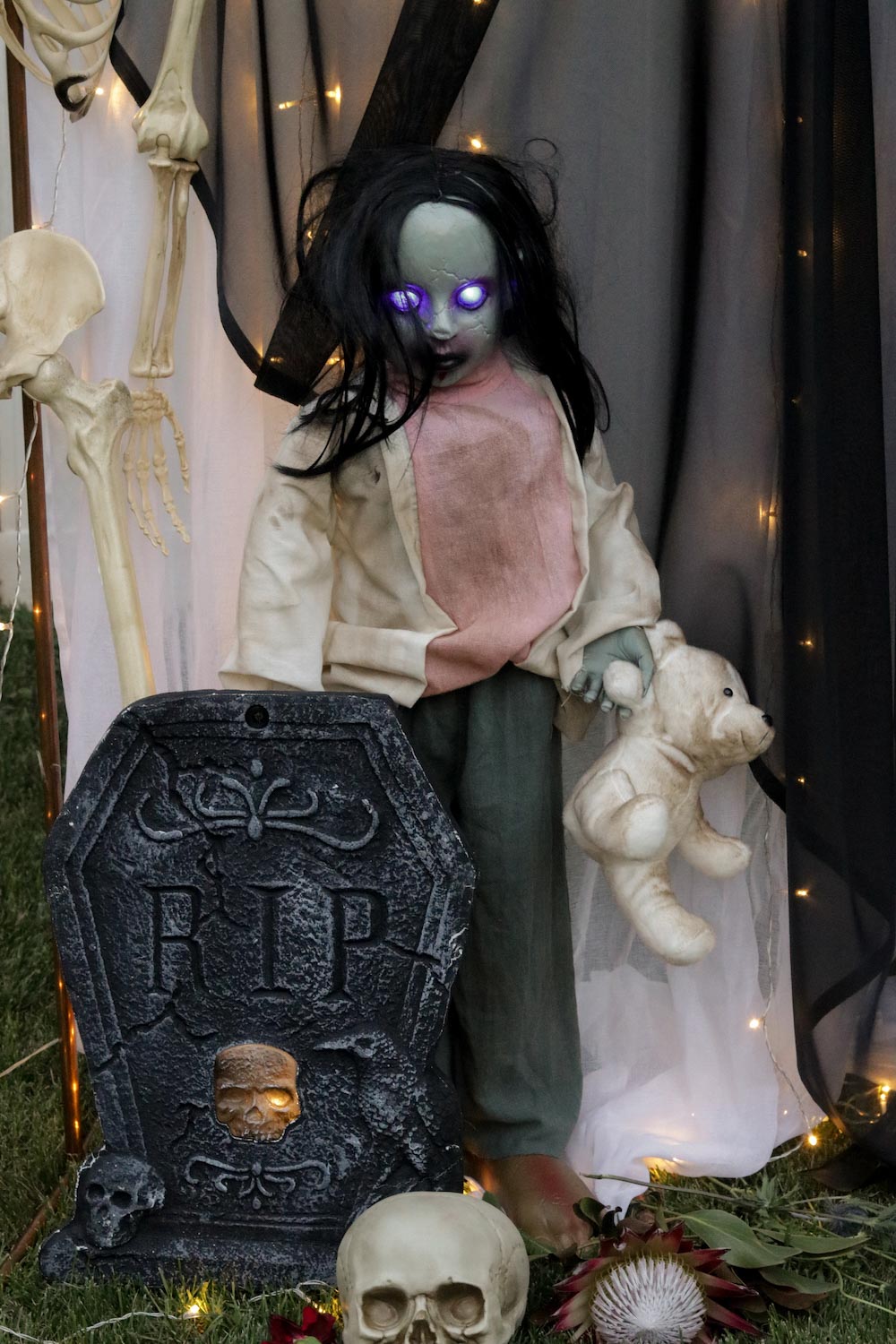 A zombie girl with glowing eyes and teddy bear standing in front of a tombstone.