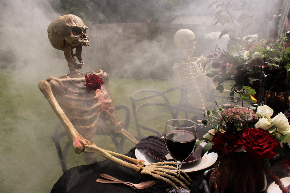 A skeleton sitting at a foggy table decorated with flowers and black linens.