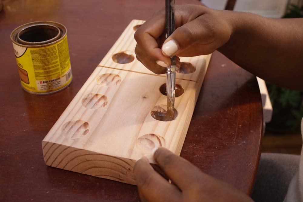 A person hand painting dark brown wood stain into carved dominoe holes on light block of wood. A person hand painting dark brown wood stain into carved dominoe holes on light block of wood.