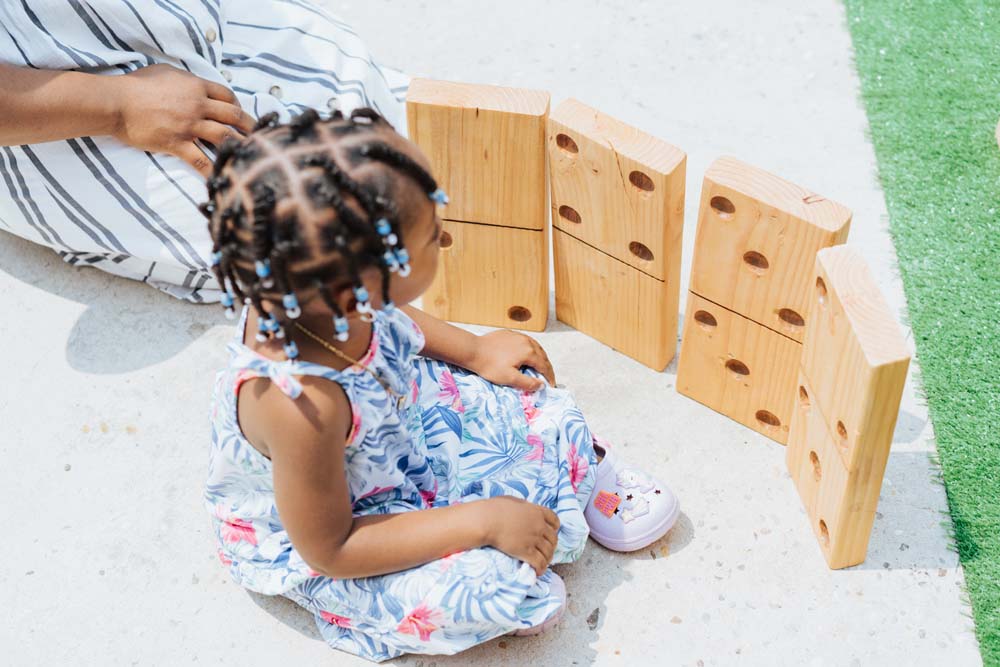 Little girl sitting in floral sundress next to kneeling women and stack of large wooden DIY dominoes. Little girl sitting in floral sundress next to kneeling women and stack of large wooden DIY dominoes.