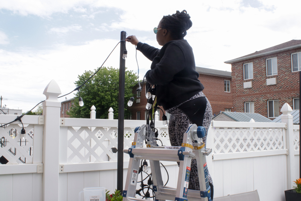 A woman standing on a ladder putting string lights on a post.