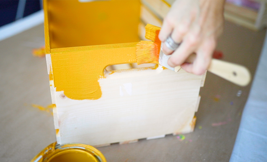 A person uses a paint brush to paint a wooden crate orange.