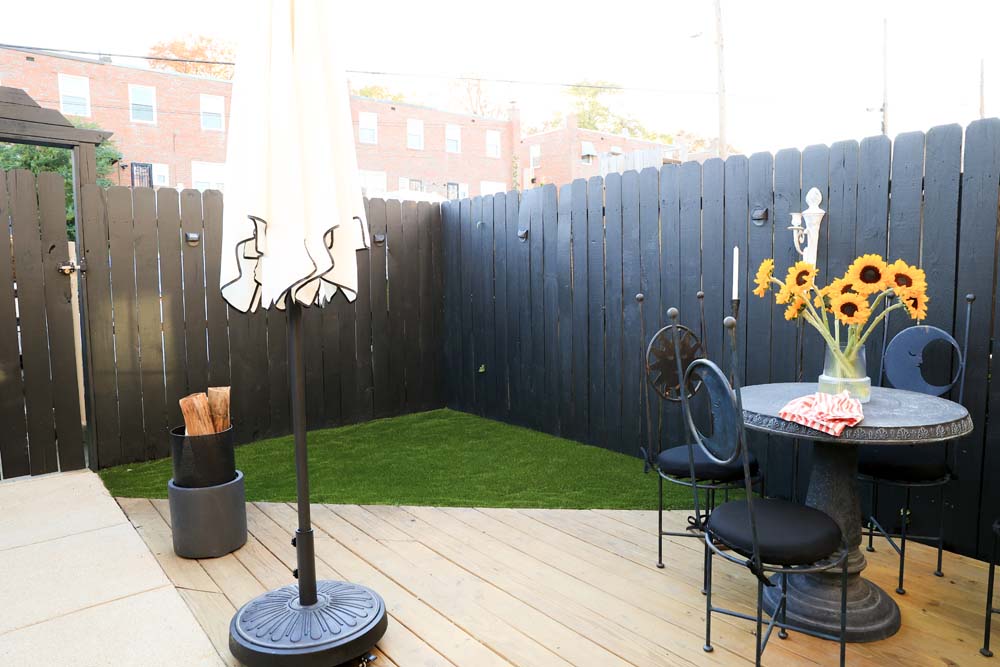 Corner of outdoor patio covered with green artificial turf with a white outdoor umbrella, small circle table with a vase of sunflowers, and a bucket of chopped wood 