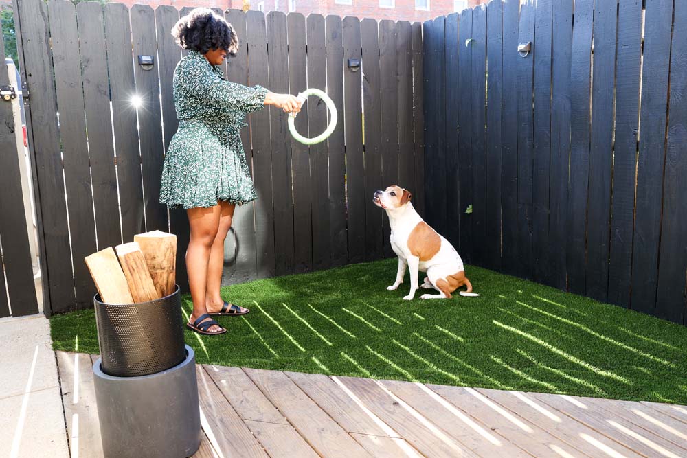 Women in a green dress standing in the corner of a outdoor patio with turf and a tan and white dog, playing with a circle rope toy. 